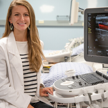 Female student wearing a lab coat sitting next to an ultrasound machine.