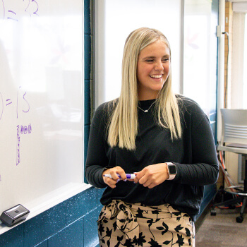 Concordia alum teaches her mathematics class, using a purple dry erase marker on a whiteboard.